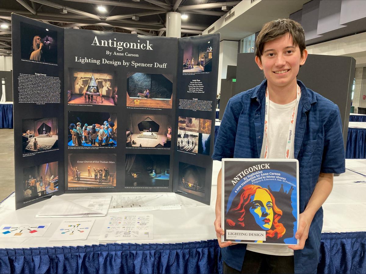 Spencer Duff poses for a photo holding with a binder and standing in front of a poster board of the play Antigonick 